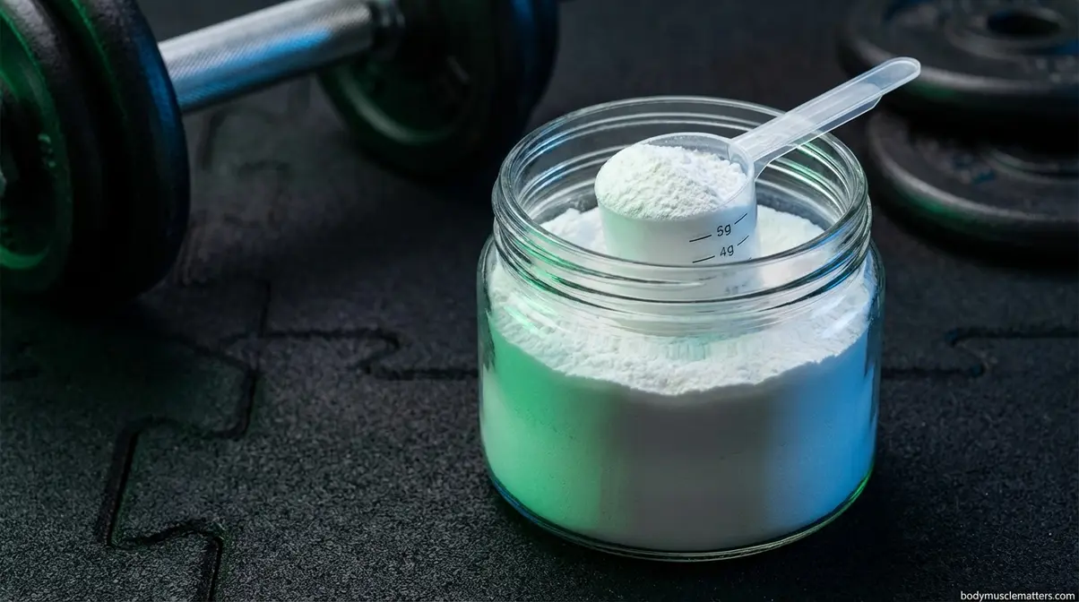 Creatine monohydrate powder with 5g measuring scoop on gym mat next to dumbbell for strength training