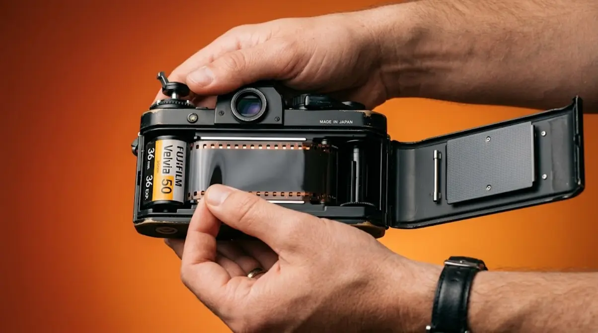 Hands loading a fresh roll of 35mm film into an open SLR camera with film leader across the channel